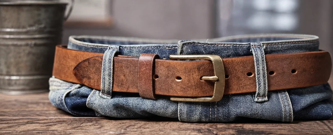 Cinto de couro masculino marrom sobre calça jeans dobrada, com fivela metálica em destaque.